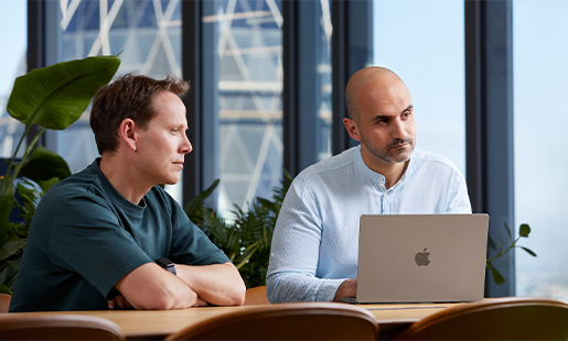 Ahmed sidder med sin MacBook foran sig ved et bord med en kollega og ser på nogen uden for billedet.