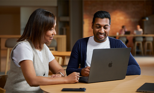 Amar i samarbejde med en kollega, mens han sidder med sin MacBook ved et bord i et fællesområde.