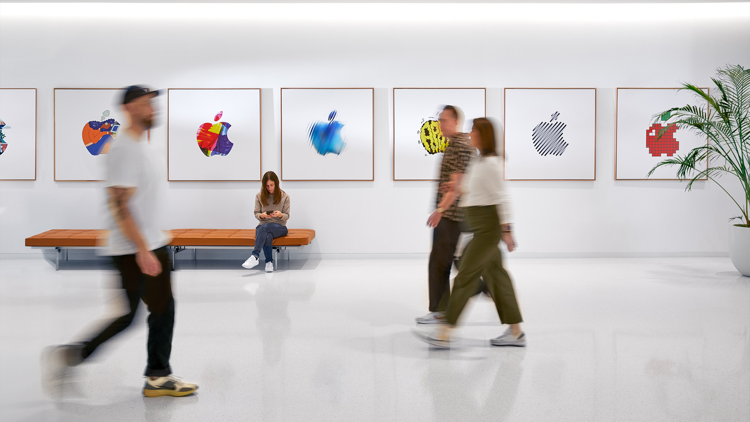 Three Apple colleagues walking past an interior wall with colourful Apple logos and a fourth employee sitting on a bench.