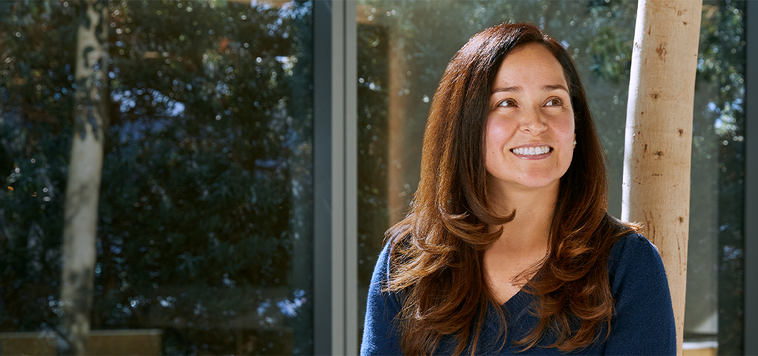 Montse smiling and sitting by a large sunny window, with trees in the background.