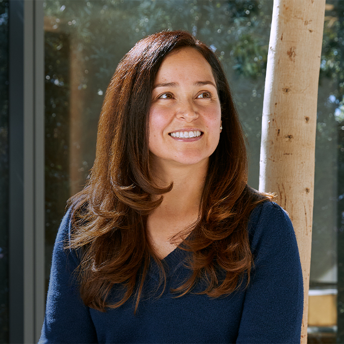 Montse smiling and sitting by a large sunny window, with trees in the background.