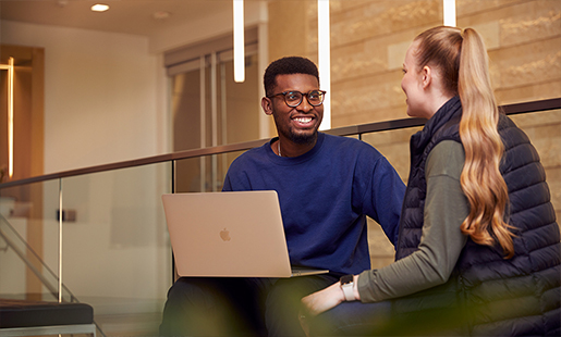 Chidi sidder med en MacBook i skødet og taler med en kollega.