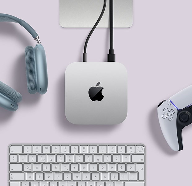 Arka bağlantı noktalarına iki adet kablonun bağlı olduğu gümüş rengi Mac mini ile yanında yer alan mavi bir AirPods Max, gümüş rengi bir Magic Keyboard ve kablosuz bir oyun kumandasının üstten görünümü