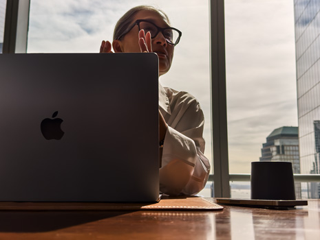 En kvinne sitter ved et bord i et høyhus. Bak henne er et stort vindu med utsikt til andre bygninger. Foran henne er en åpen MacBook.