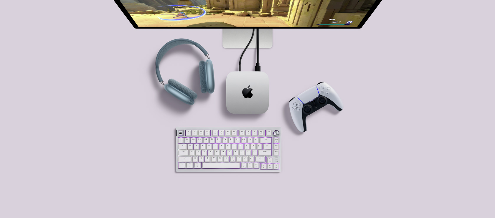 Top view of a silver Mac mini plugged into a display showing a game in active play, and next to a wireless keyboard, a gaming controller and blue AirPods Max