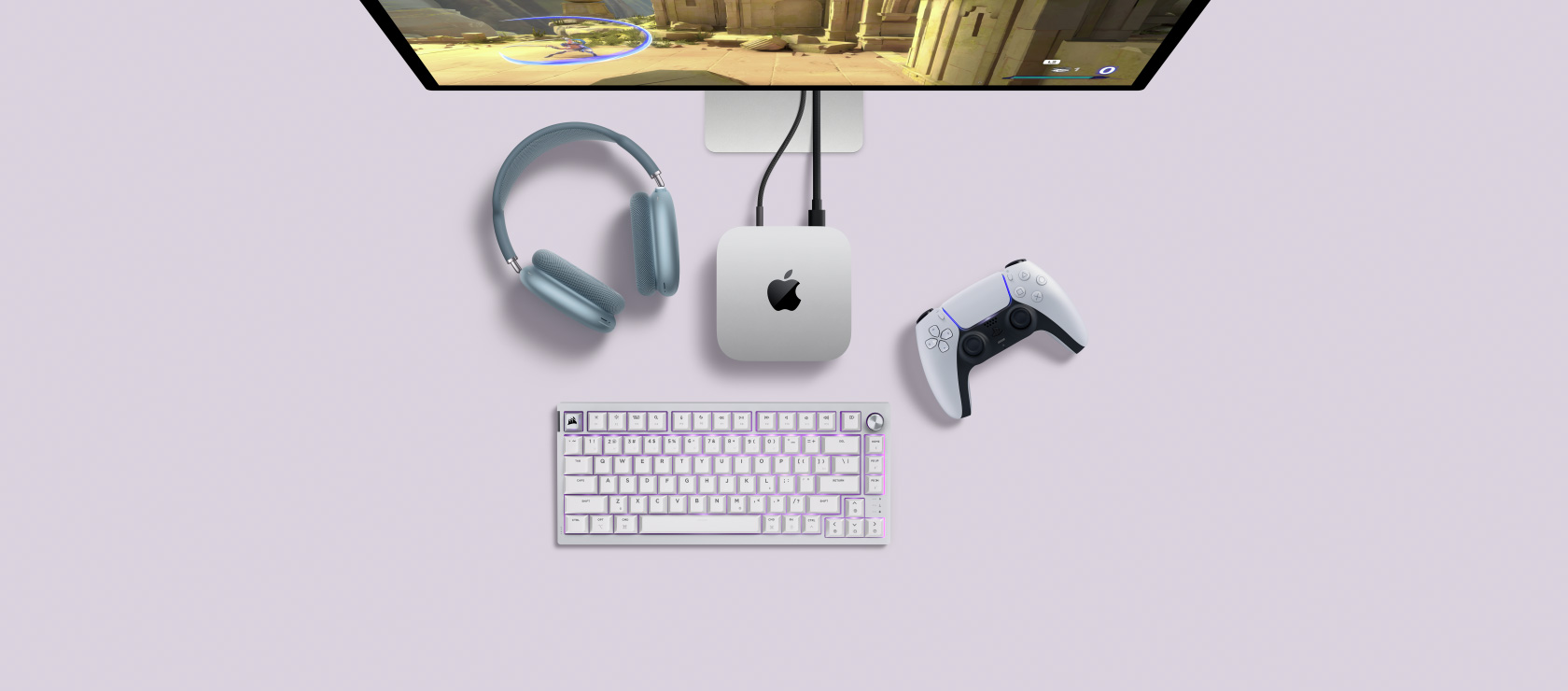 Top view of a silver Mac mini plugged into a display showing a game in active play, and next to a wireless keyboard, a gaming controller, and blue AirPods Max