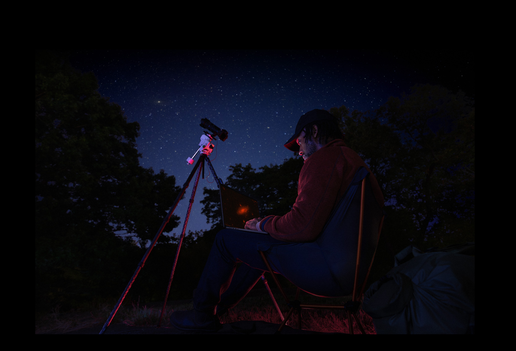 Seseorang berada di luar pada malam hari sedang menggunakan MacBook Pro di pangkuan, terhubung ke perangkat pada tripod yang mengarah ke langit penuh bintang